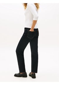 Dunkelblaue Jeans aus Denim mit geradem Schnitt, mit Gesäßtaschen und kontrastierenden Nähten. Ein lockeres weißes Oberteil und schwarze Slipper runden den Look ab.