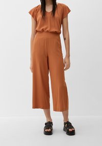 Haut couleur rouille à manches courtes avec col en V, associé à un pantalon à jambes larges. Tenue fabriquée en tissu léger ; elle est complétée par des sandales à plateforme noires.