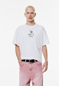 Witte t-shirt met een autoprint, gecombineerd met lichtroze spijkerbroek. Bevat een zwarte riem en gelaagde zilveren sieraden. Eenvoudige achtergrond.