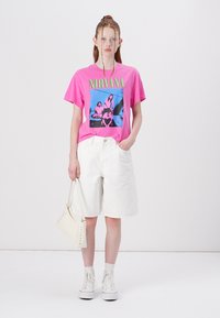 Lysende rosa t-skjorte med "NIRVANA" grafikk, parret med hvite denimshorts, hvite høyhælte sneakers og en liten hvit veske.