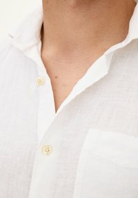 Camicia bianca in lino con colletto, una tasca sul petto e chiusura a bottoni con una sottile lucentezza. Tessuto strutturato con un aspetto leggero.