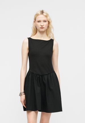 Femeie cu păr lung blond, purtând o rochie neagră fără mâneci, cu o fustă adunată, stând în picioare în fața unui fundal simplu și luminos.