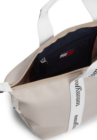 Μπεζ υφασμάτινη τσάντα με λευκές λουρίδες ώμου, εσωτερική τσέπη με φερμουάρ και εμφανές λογότυπο Tommy Jeans. Ομαλή υφή και casual σχεδιασμός.