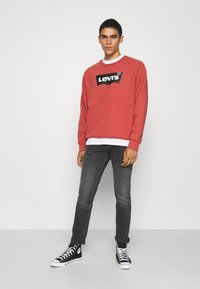 Felpa rossa a girocollo con logo Levi's nero, indossata su una maglietta bianca, abbinata a jeans scuri e sneakers alte nere.