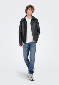 Giacca nera in ecopelle con collo a risvolto, indossata sopra una T-shirt bianca, abbinata a jeans azzurri e sneakers grigie.