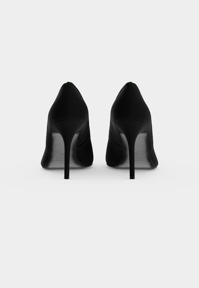 Paire d'escarpins noirs vus de dos, avec des talons aiguilles hauts et fins sur un fond gris clair uni.