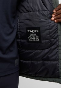 Texapore Core jacket interior, dark quilted lining, waterproof label, stitching details, green trim, and logo tag on fabric.