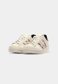 Cream leather sneakers with a textured toe cap, black heel, and brown striped accents. Features flat laces and padded collar for comfort.