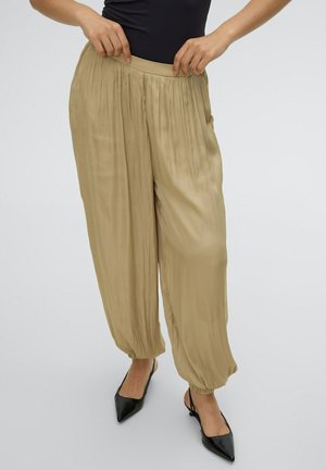 Mujer ajustándose unos pantalones beige plisados y sueltos con tobillos elásticos, llevando zapatos slingback negros de punta puntiaguda y una blusa negra sobre un fondo blanco.