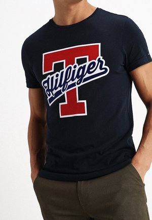 Muž v námořnickém modrém tričku s velkým červeným "T" a bílým textem "Hilfiger" na hrudi, v kombinaci s olivově zelenými kalhotami.