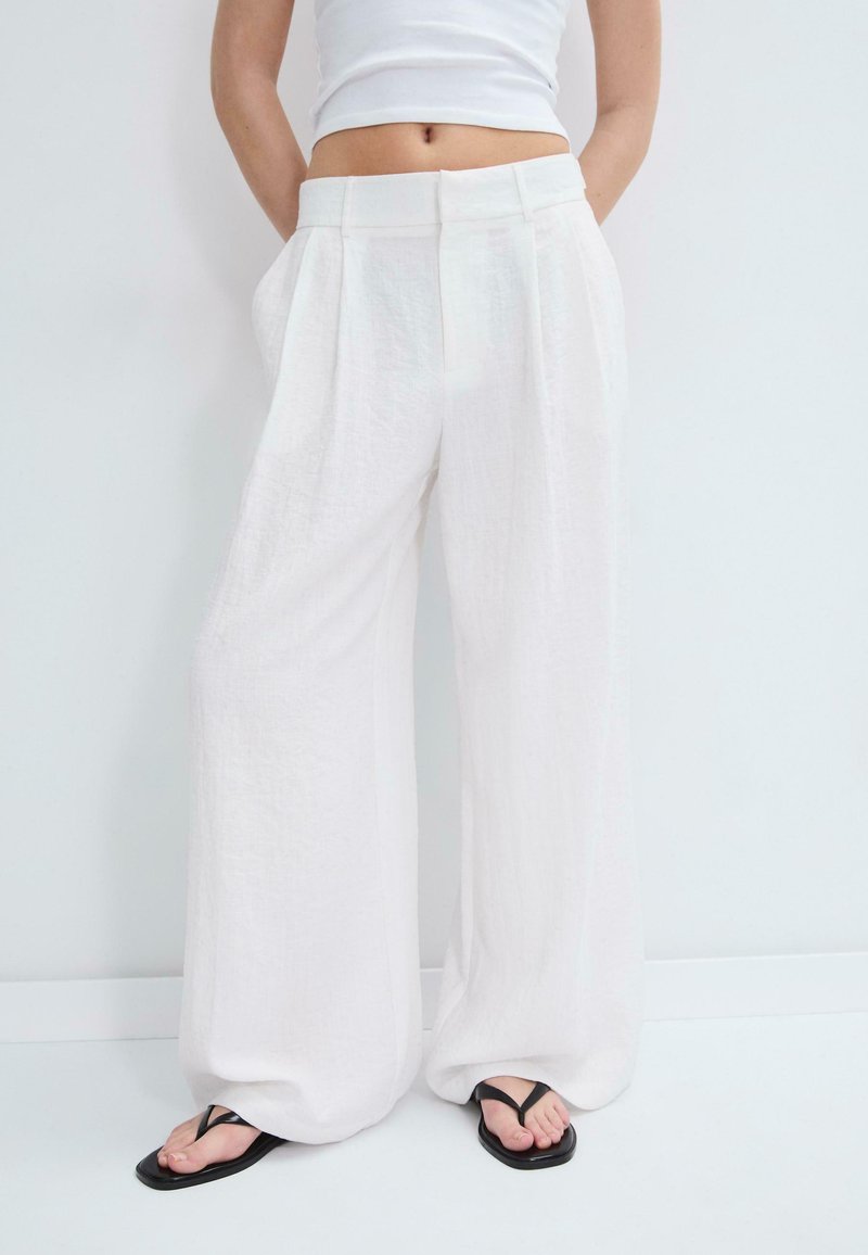 Personne portant un pantalon ample en lin blanc, un débardeur court blanc et des tongs noires à bout carré, debout devant un fond blanc.