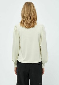 Maglietta a maniche lunghe color crema con spalle a sbuffo, texture liscia, orlo arrotondato e design minimalista, indossata con pantaloni neri.