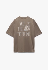 Brun oversized t-shirt med korte ærmer. Hvid tekst på ryggen siger "VI ER FREMTIDEN" med en pil og en blomst grafik. Blød tekstur.