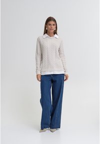 Maglione crema a trecce con maniche lunghe indossato sopra una camicia bianca con colletto, abbinato a jeans in denim blu a gamba larga e scarpe beige con punta appuntita.