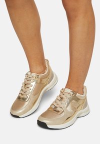Las zapatillas metálicas doradas tienen una textura suave, forma redondeada y cordones suaves. Los acentos incluyen paneles laterales y una suela acolchada.