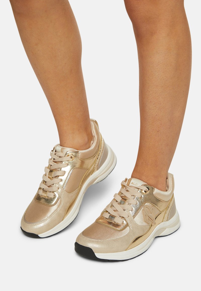 Las zapatillas metálicas doradas tienen una textura suave, forma redondeada y cordones suaves. Los acentos incluyen paneles laterales y una suela acolchada.