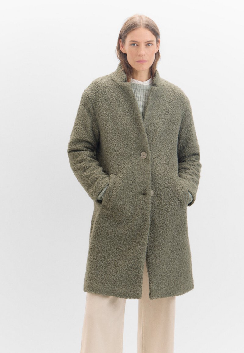Olivgrüner, strukturierten Mantel aus plüschigem Material, mit einem Reverskragen, zwei frontalen Taschen und einem einzelnen Knopfverschluss.