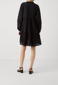 Vestido negro de ojal con mangas largas, cuello redondo, dobladillo en capas y un sutil patrón de recortes. Lleva unas sandalias negras de tacón cuadrado.