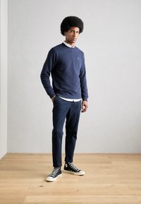 Marineblauwe trui met een ronde hals, gecombineerd met donkerblauwe broek en zwarte hoge sneakers. Glad materiaal, ontspannen pasvorm, minimale branding.