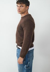 Brauner Strickpullover mit rundem Halsausschnitt und gerippten Bündchen, kombiniert mit blauen lässigen Jeans mit Seitentaschen und einem Gürtel.