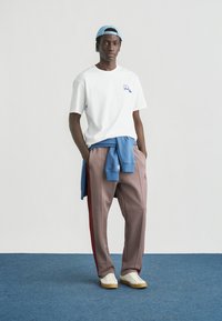 T-shirt en coton blanc avec logo bleu, pantalon à carreaux rouges et bleus, pull bleu clair attaché à la taille, baskets blanches avec semelle crème.