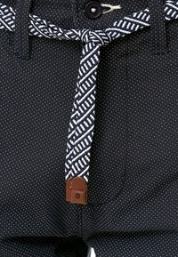 Gros plan sur un pantalon noir à petits points blancs et une ceinture tressée noire et blanche nouée avec des extrémités en cuir marron.