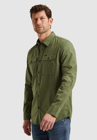 Camicia a maniche lunghe verde oliva con chiusura a bottoni, due tasche sul petto e una vestibilità comoda. Realizzata in cotone con una texture liscia.