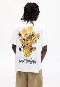 Weißes, übergroßes T-Shirt mit einem lebhaften gelben Sonnenblumenmuster und dem Text "Vincent Van Gogh" in einer strukturierten Optik. Kurze Ärmel, lockere Passform.