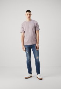 Camiseta de crew-neck en color púrpura claro combinada con jeans azules desgastados y zapatillas blancas. Los jeans tienen desgarros cerca de las rodillas. Diseño simple.