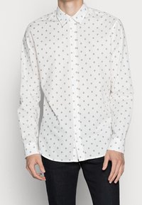 Camicia bianca con bottoni caratterizzata da un piccolo motivo circolare ripetuto. Realizzata in tessuto liscio con colletto classico e allacciatura con bottoni.