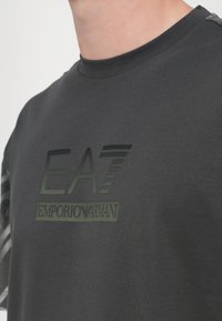 Tamsiai pilkas trumpomis rankovėmis marškinėliai su matinio žalia EA7 Emporio Armani logotipu. Yra apvali iškirptė ir lygaus audinio tekstūra.