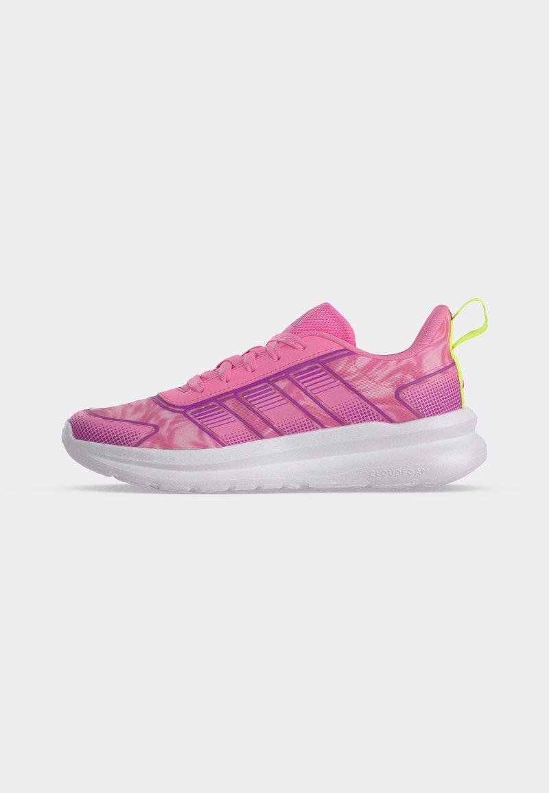 Zapatilla deportiva rosa con rayas moradas, suela blanca acolchada y tira de sujeción en neón amarillo en el talón, mostrada en perfil lateral sobre fondo blanco.