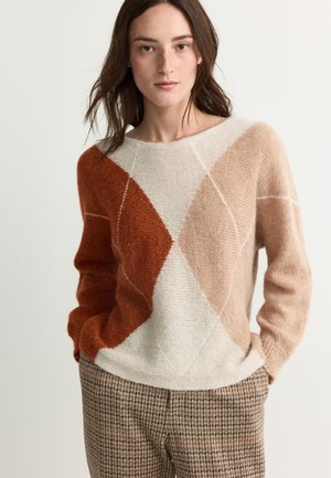 Frau trägt einen Pullover mit geometrischem Muster in Creme, Rost und Beige sowie karierte braune Hosen, steht mit den Händen in den Taschen.