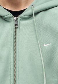 Φωτεινό πράσινο φούτερ με λευκό λογότυπο Nike στο στήθος και κορδόνι, φορεμένο πάνω από μαύρο πουκάμισο.