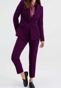 Lila kostym med en åtsittande kavaj och matchande smala byxor. Kavajen har en knapp och två framfickor.