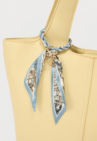 Φωτεινή κίτρινη τσάντα με πλεκτό μπλε και λευκό φλοράλ μαντίλι δεμένο γύρω από τη λαβή, διακοσμημένη με ένα μικρό χρυσό στρογγυλό γούρι.