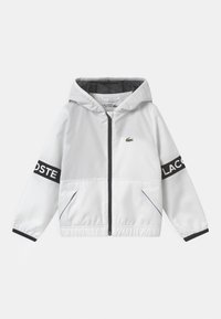 White windbreaker jacket with a hood, black accents, Lacoste logo on sleeves, lined interior, elastic cuffs, and zip closure.