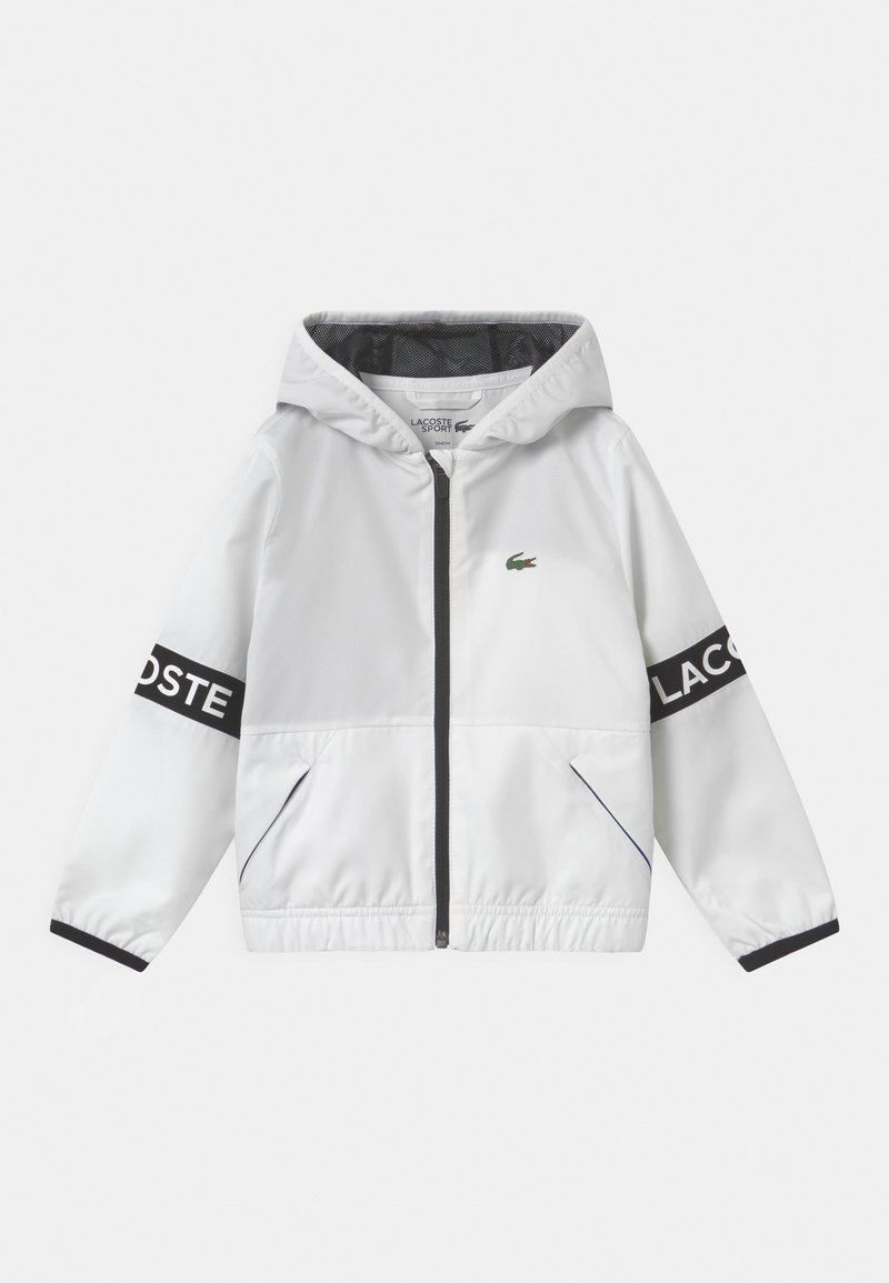 White windbreaker jacket with a hood, black accents, Lacoste logo on sleeves, lined interior, elastic cuffs, and zip closure.