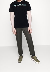 Černé tričko s grafikou "TOM TAILOR", olivově zelené kalhoty a olivové tenisky s bílými akcenty, s kulatým výstřihem a krátkými rukávy.