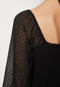 Svart klänning med texturerade, prickiga genomskinliga ärmar och smockad bodice, med en bred V-ringning och avslappnad passform.