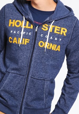 Osoba ubrana w niebieską bluzę z kapturem na suwak z żółtym napisem „Hollister California” i białymi sznurkami, pod którą ma ciemnoniebieską koszulkę.