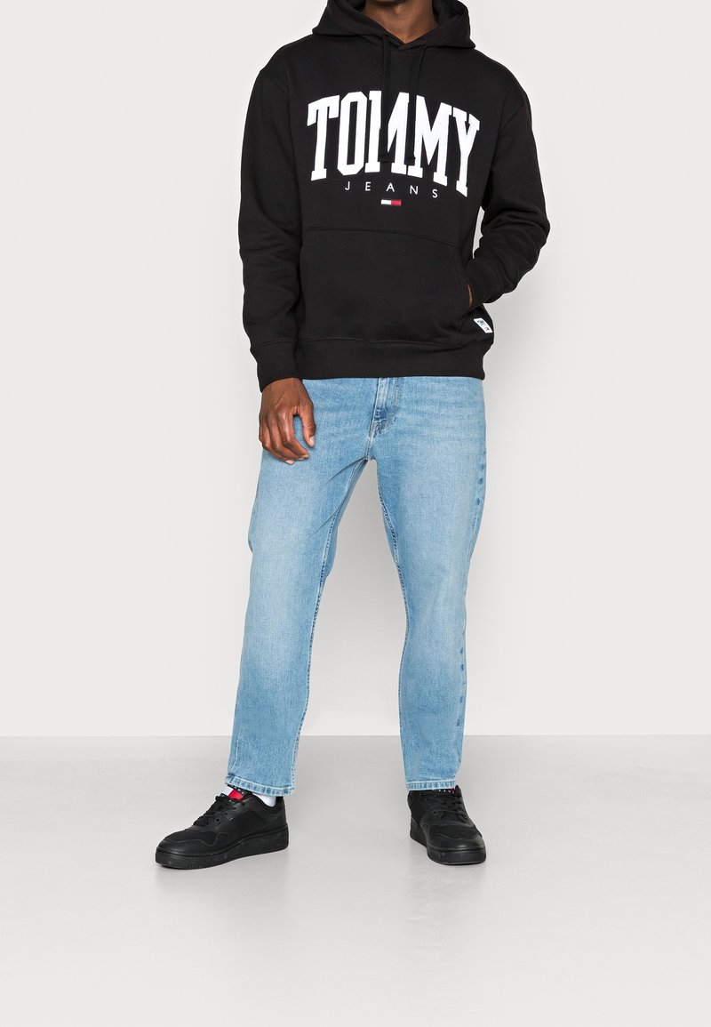 Personne portant un sweat à capuche noir Tommy Jeans, un jean bleu clair, des baskets noires, debout devant un fond clair uni avec une main dans la poche.