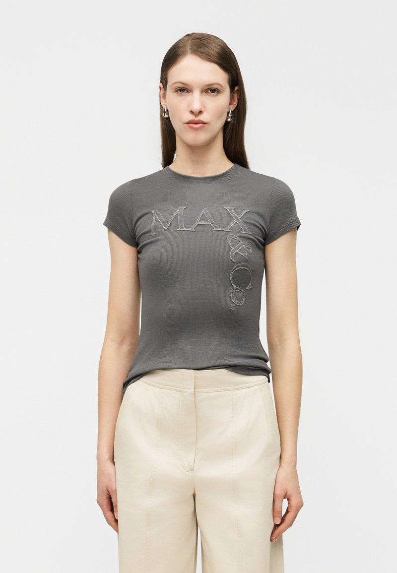 Γυναίκα με μακριά καστανά μαλλιά, φοράει εφαρμοστή γκρι μπλούζα με ριμπ ύφανση και την επιγραφή "MAX & CO." και μπεζ παντελόνι ψηλόμεσο με φαρδιά μπατζάκια.