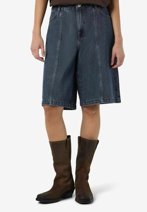 Persoon draagt donkere denim shorts tot op de knie en bruine leren laarzen tot halverwege de kuit, staand tegen een effen achtergrond.