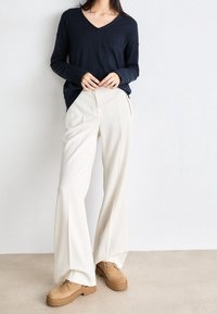 Kobieta w luźnym granatowym swetrze w serek, szerokich białych spodniach i brązowych zamszowych butach na sznurówki stoi przy gładkiej białej ścianie.