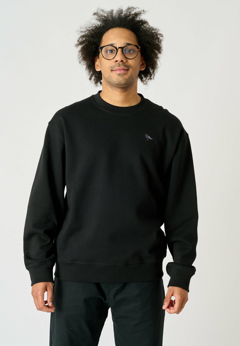 Schwarzer Sweatshirt mit geripptem Textur, rundem Halsausschnitt und langen Ärmeln. Besitzt ein kleines graues Vogel-Logo auf der Brust. Einfaches Design.