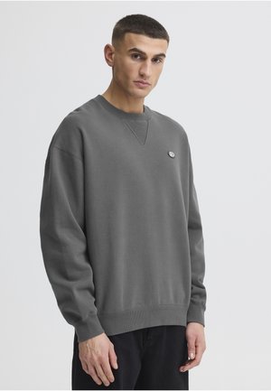 Junger Mann mit kurzen dunklen Haaren, der einen lockeren dunkelgrauen Pullover und schwarze Hose vor einem schlichten hellen Hintergrund trägt.