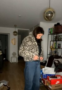 Egy nő, aki egy leopárdmintás dzsekit és kék farmert visel, gombokat tűz egy nappaliban, ahol polcok és egymásra rakott cipősdobozok vannak a közelben.