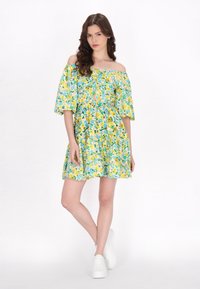 Off-the-shoulder jurk met een bloemmotief in geel, groen en blauw. Korte mouwen, getailleerde lijfje en een gelaagde rok. Witte sneakers.