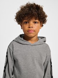 Barn med krøllet hår iført en grå hættetrøje med sorte mærkede striber på ærmerne, der ser direkte ind i kameraet mod en ensfarvet baggrund.